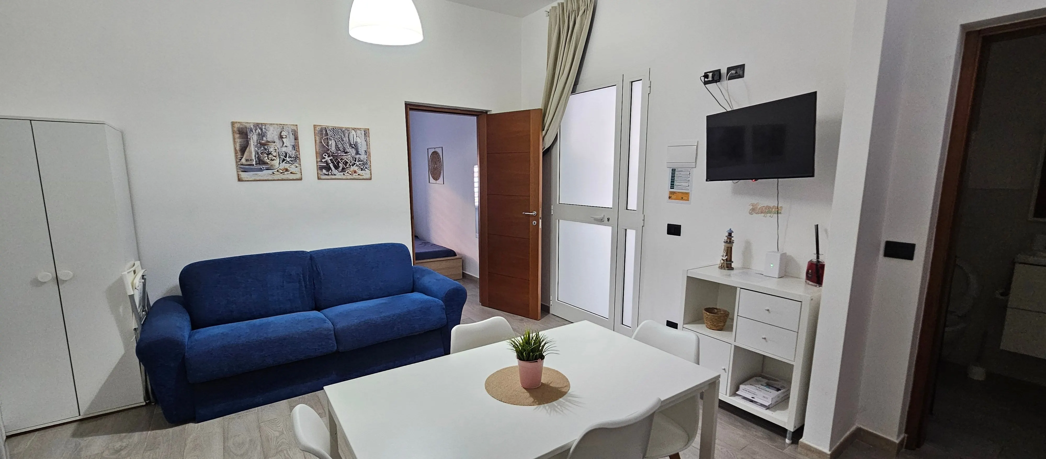 Pièce de vie de l'appartement : canapé bleu, table blanche, TV et portes intérieures.